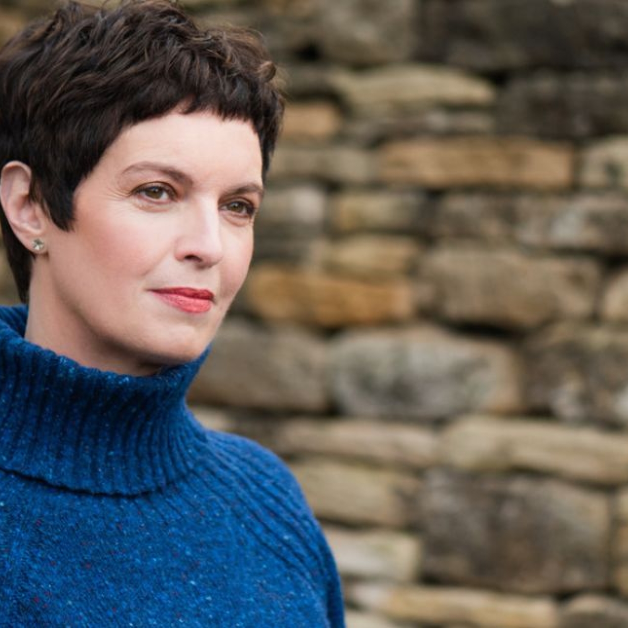 A colour portrait of Louise Tickle in front of a stone wall. Tickle is a white woman with short dark hair wearing a blue polo necked jumper. She faces at angle to the camera.